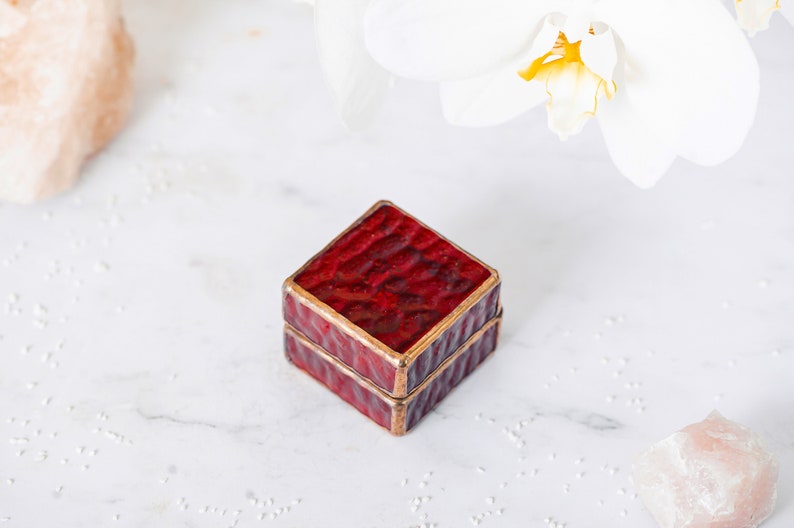 May include: A small, square, red ring box with a gold trim. The box is closed and sitting on a white surface.