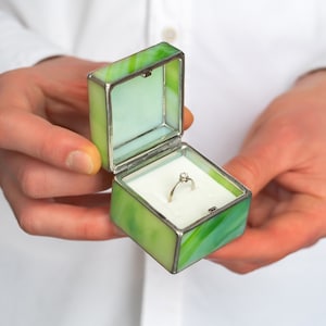May include: A green glass ring box with a silver trim and a white interior. A diamond ring is inside the box.