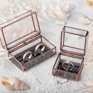 May include: Two glass ring boxes with copper trim. One box holds two gold wedding bands, the other holds a single diamond engagement ring.