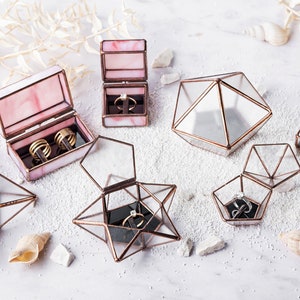 May include: Assortment of glass ring boxes in various geometric shapes, including a star, pyramid, and rectangle. Some boxes are open and show a black velvet interior with rings inside. The boxes are made of clear glass with copper accents.