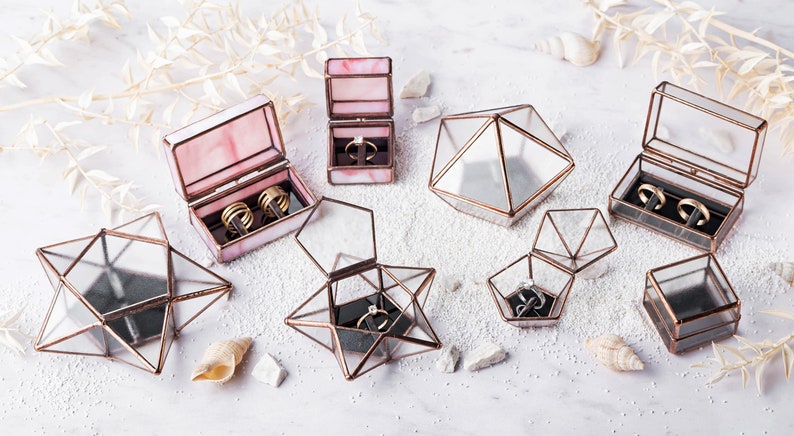 May include: Assortment of glass ring boxes in various geometric shapes, including a star, pyramid, and rectangle. Some boxes are open and show a black velvet interior with rings inside. The boxes are made of clear glass with copper accents.