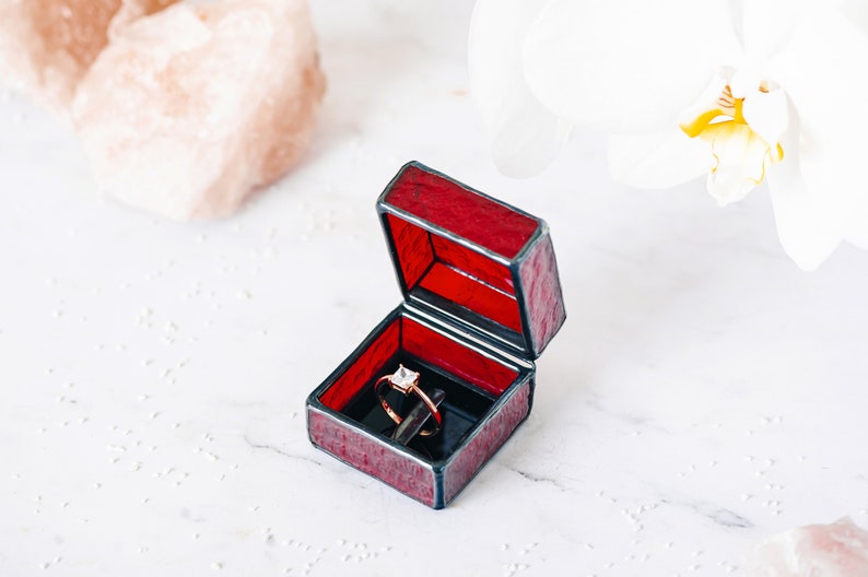 May include: A red stained glass ring box with a gold ring inside. The box is open and the ring is visible. The box is sitting on a white surface.