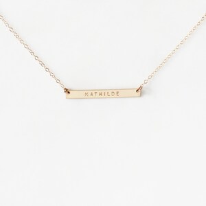 May include: A gold chain necklace with a thin, horizontal bar pendant engraved with the name 'MATHILDE'.