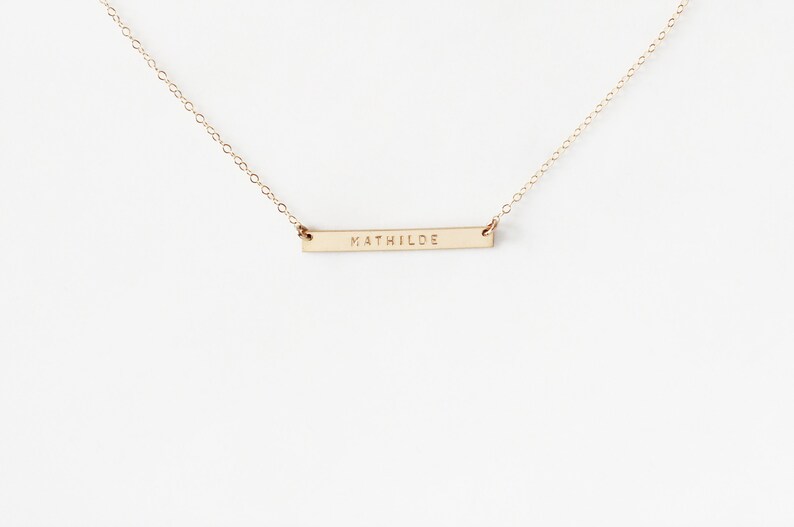 May include: A gold chain necklace with a thin, horizontal bar pendant engraved with the name 'MATHILDE'.
