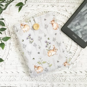 May include: A pale grey Kindle cover featuring a pattern of brown rabbits, white flowers, and green leaves. The cover has a wooden button fastening and a grey polka dot ribbon. A Kindle e-reader is visible.