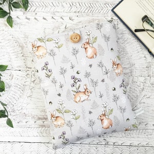 May include: A light grey book sleeve with a repeating pattern of watercolour rabbits, flowers, and foliage. The sleeve is fastened with a wooden button. A book and glasses are in the background.