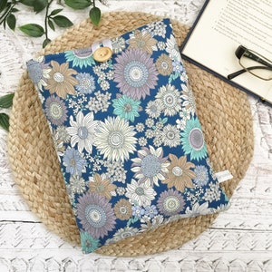 May include: A navy blue book cover with a floral design of sunflowers in white, lavender, and brown. It is fastened with a wooden button and white ribbon. The cover rests on a woven mat. The book cover is designed to protect a book.