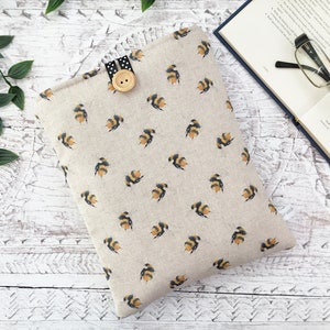May include: A beige fabric book sleeve featuring a pattern of black and yellow bumblebees. It has a black and white polka dot tab closure with a wooden button. A book and glasses are in the background.