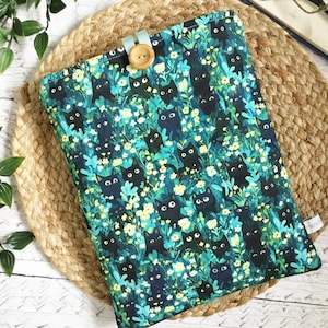 May include: A rectangular book sleeve with a whimsical cat pattern in teal, green, and yellow. The fabric features black cat illustrations amidst floral elements. A wooden button and teal loop secure the sleeve. The sleeve is approximately 25 cm x 20 cm.