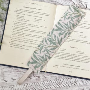 May include: An open book with a fabric bookmark featuring a white background and a green leaf design. The book displays text about cleaning boots and shoes. The book has a dark blue cover and is resting on a light-colored surface.
