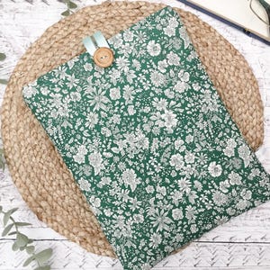 May include: A rectangular fabric sleeve featuring a dark green background with a dense white floral pattern. The sleeve is secured at the top centre with a light blue fabric loop and a single round, light brown wooden button closure.