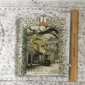 K&ouml;nnte beinhalten: Eine Stoffh&uuml;lle f&uuml;r B&uuml;cher mit gr&uuml;n-wei&szlig;em Blattmuster und einem Holzknopfverschluss. Die H&uuml;lle enth&auml;lt das Buch Fried Green Tomatoes at the Whistle Stop Cafe von Fannie Flagg. Ein Holzlineal zeigt eine H&ouml;he von 23 cm an.