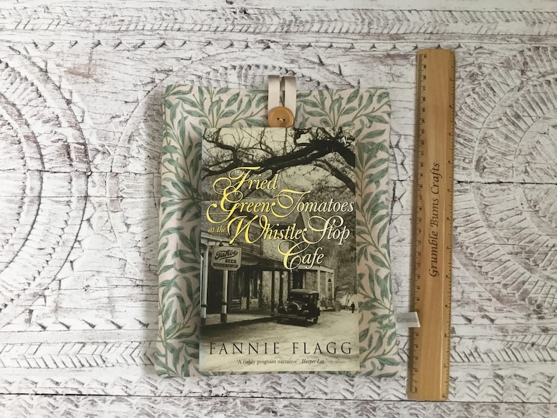 K&ouml;nnte beinhalten: Eine Stoffh&uuml;lle f&uuml;r B&uuml;cher mit gr&uuml;n-wei&szlig;em Blattmuster und einem Holzknopfverschluss. Die H&uuml;lle enth&auml;lt das Buch Fried Green Tomatoes at the Whistle Stop Cafe von Fannie Flagg. Ein Holzlineal zeigt eine H&ouml;he von 23 cm an.