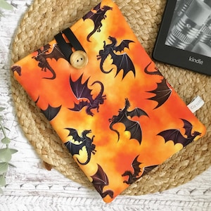 May include: An orange and yellow fabric Kindle cover with a repeating pattern of black dragons. The cover has a black strap and wooden button closure. A Kindle e-reader is visible.
