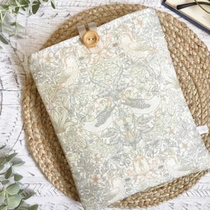 May include: A patterned book cover featuring birds and floral motifs in grey, beige, and green hues. It fastens with a wooden button and a beige strap. The cover is placed on a woven mat, suitable for a book.