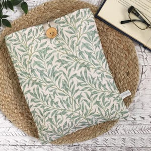 May include: A book sleeve with a green and off-white leaf pattern, secured with a wooden button. It rests on a woven mat, with a book and glasses nearby. The design is reminiscent of a botanical print.
