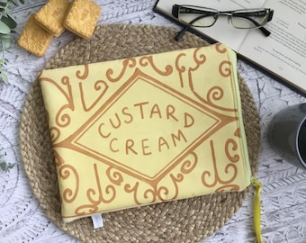 Może przedstawiać: Prostokątna kosmetyczka zapinana na zamek w kolorze jasnego żółtego z brązowym nadrukiem graficznym przedstawiającym herbatnik custard cream, ozdobiona ornamentami i napisem CUSTARD CREAM wyśrodkowanym w rombie na przedniej stronie.