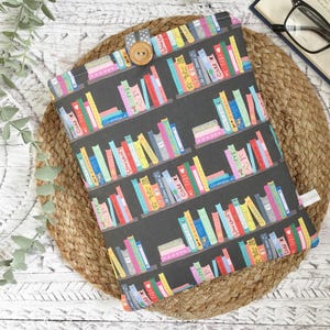 May include: A book cover with a black background and a colourful bookshelf pattern. It fastens with a wooden button and a grey fabric loop. The cover is placed on a woven mat, with a book and glasses visible.