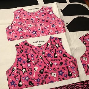 May include: Fabric pieces for a doll vest, sleeves, and front, featuring a pink background with white and purple stars. The vest back has a pattern of guitars, musical notes, and the text "Rock Star". The words "LET'S PLAY DOLLS" are printed on the fabric.