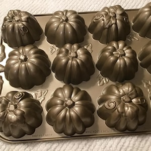 May include: A bronze-colored pumpkin-shaped baking pan with 12 individual molds. The pan has a decorative leaf design on the side and is ideal for making festive fall treats. Each mold is approximately 3 inches in diameter.