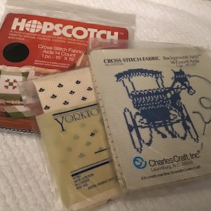 May include: Three packages of cross stitch fabric including a red Hopscotch 14 count Aida 15 inch by 15 inch piece, a Yorktown ecru 14 count 14 inch square piece, and a Charles Craft 14 count Aida fabric featuring a blue horse-drawn carriage design.
