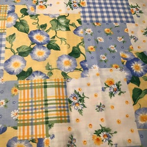 May include: A patchwork textile featuring various floral and gingham patterns. The design incorporates blue morning glories, daisies, and yellow and green plaid squares. The overall color scheme includes blue, yellow, white, and green.