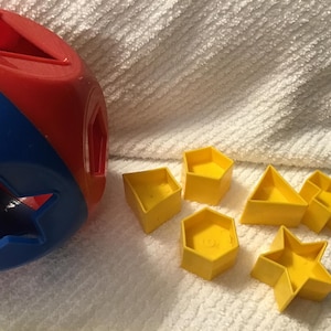 May include: A red and blue shape sorter toy with various yellow geometric shapes. The shapes include a cube, triangle, hexagon, star, and a cross-like shape. The toy is designed for early childhood development.