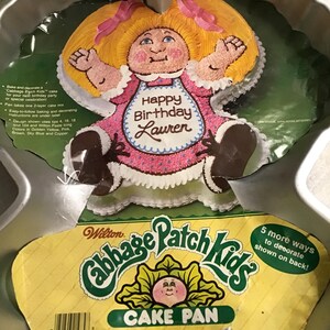May include: A Cabbage Patch Kids cake pan with a cake shaped like a doll wearing a pink dress and apron that says "Happy Birthday Lauren". The pan is silver and has a yellow label that says "Cabbage Patch Kids Cake Pan".