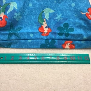 May include: A blue tote bag featuring a repeating pattern of a mermaid with red hair and a green tail, along with starfish and floral designs. A green ruler with inch measurements is at the bottom.