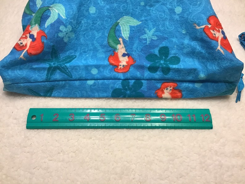 May include: A blue tote bag featuring a repeating pattern of a mermaid with red hair and a green tail, along with starfish and floral designs. A green ruler with inch measurements is at the bottom.