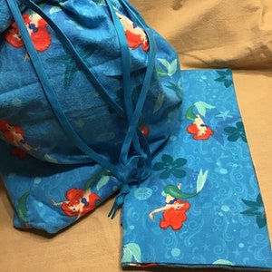 May include: A blue drawstring bag and a matching rectangular item, both featuring a mermaid print. The mermaid has red hair and a green tail, set against a background of blue and green aquatic elements.