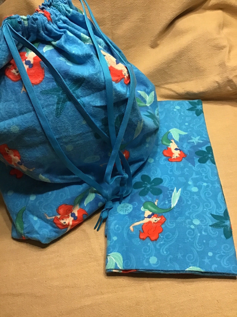 May include: A blue drawstring bag and a matching rectangular item, both featuring a mermaid print. The mermaid has red hair and a green tail, set against a background of blue and green aquatic elements.