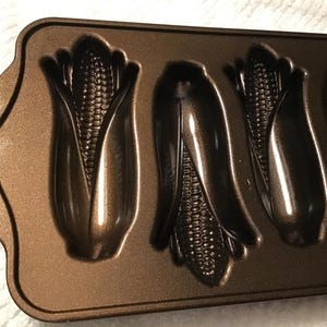 May include: A bronze-colored, non-stick cornbread pan with five corn cob-shaped molds. The pan has a rectangular shape with a curved handle on the left side. The molds are detailed with kernels and husks.