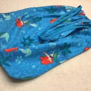 May include: A drawstring bag featuring a vibrant blue fabric with a mermaid pattern. The design includes mermaids with red hair and green tails, along with starfish and floral accents. The bag has a blue drawstring closure.