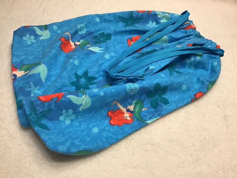 May include: A drawstring bag featuring a vibrant blue fabric with a mermaid pattern. The design includes mermaids with red hair and green tails, along with starfish and floral accents. The bag has a blue drawstring closure.