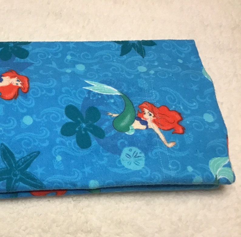 May include: Folded blue fabric featuring a mermaid design with red hair and a green tail. The fabric has a pattern of swirling water, starfish, and flowers. The mermaid is swimming in the water. The fabric is folded over.
