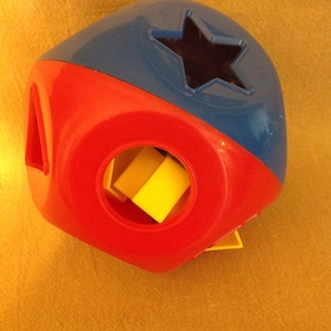 May include: A colorful shape sorter toy in red and blue with a yellow shape inside. The toy has a star-shaped hole and other geometric openings. The toy is designed for early childhood development.