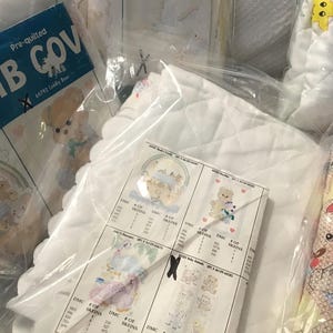 May include: A collection of pre-quilted crib covers and related items, packaged in clear plastic. The packaging includes a blue and white label that reads "CRIB COV" and features a teddy bear illustration. Other items include white quilted fabric and printed patterns.