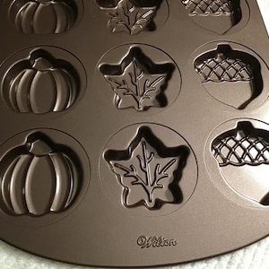 May include: A brown metal baking pan with pumpkin, leaf, and acorn shapes. The pan has a non-stick surface and is designed for making fall-themed treats. The brand name "Wilton" is visible on the pan.