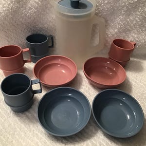 May include: A vintage plastic pitcher with a blue lid, surrounded by matching pink and blue cups and bowls. The set includes a pitcher, two bowls, two mugs, and two smaller cups, all in a retro style.