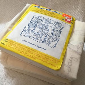 May include: A stack of off-white fabric squares topped with a yellow and white Aunt Martha's hot iron transfer packet. The packet features blue line art illustrations of roosters and the text 3753 Barnyard Happenings.