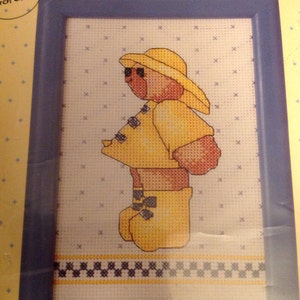 May include: A cross-stitch pattern featuring a yellow bear wearing a yellow raincoat and yellow rain boots. The bear is standing on a white background with a blue and white checkered border.