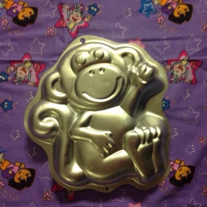 May include: A silver monkey-shaped cake pan. The monkey is sitting with its arms crossed and has a big smile.