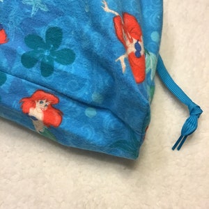 May include: A blue drawstring bag featuring a pattern of Ariel from The Little Mermaid, flowers, and aquatic designs. The bag has a blue drawstring closure. The fabric is a soft, textured material.