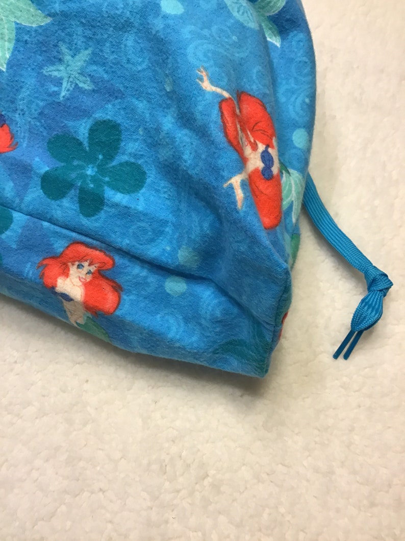 May include: A blue drawstring bag featuring a pattern of Ariel from The Little Mermaid, flowers, and aquatic designs. The bag has a blue drawstring closure. The fabric is a soft, textured material.