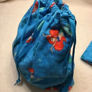 May include: A blue drawstring bag featuring a repeating pattern of a red-haired mermaid on a blue background. The bag is gathered at the top with a blue drawstring closure.
