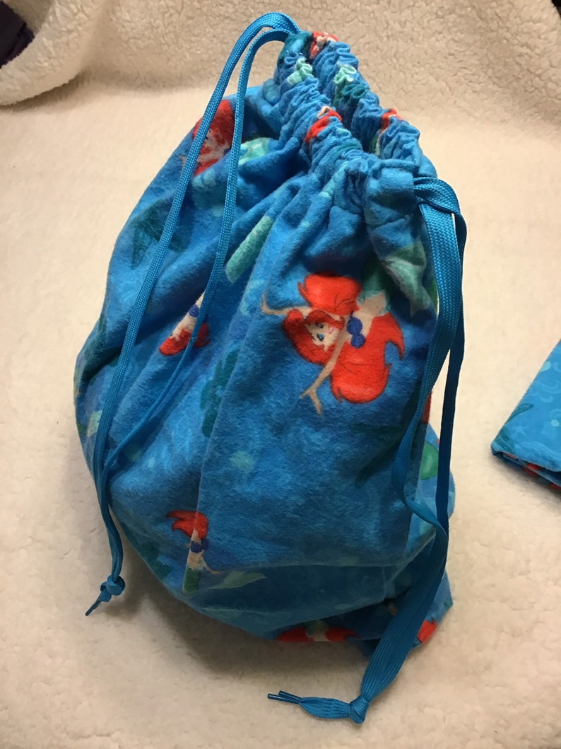 May include: A blue drawstring bag featuring a repeating pattern of a red-haired mermaid on a blue background. The bag is gathered at the top with a blue drawstring closure.