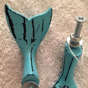 May include: Two turquoise mermaid tail cabinet knobs with distressed black accents. Each knob has a metal bolt, washer, and nut for installation. The knobs are designed for furniture or decorative use.
