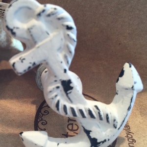 May include: White anchor-shaped decorative hook with distressed black accents. The hook has a nautical theme, featuring a rope-like design on the top portion. It is mounted on a metal base, suitable for hanging items. The hook is likely made of cast iron.