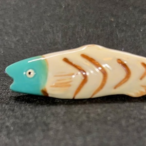 May include: A ceramic fish figurine with a turquoise head, cream-colored body, and brown markings. The fish has a detailed design with a small eye and a curved tail. The figurine is a decorative item.